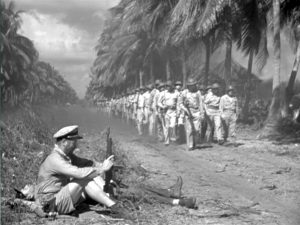 This image is a black and white frame still from the film "They Were Expendable." On the left, it shows two tired officers resting by the side of a road. On the right, a platoon of soldiers marches down the road in two rows toward the camera flanked by a row of palm trees.