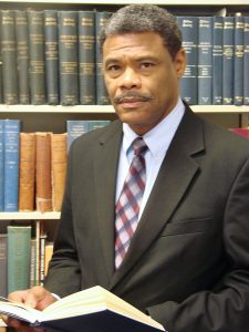 Portrait image of William Chester Jordan wearing a suit and tie standing in front of shelves of books holding an open book but looking at the camera