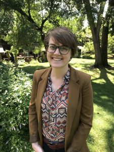 Portrait image of Rebecca Moorman standing outdoors in front of trees. Moorman has short hair, is smiling, and wears a patterned blouse under a brown jacket and dark glasses