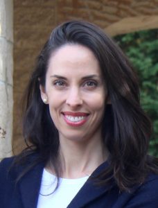 Portrait image of Gloria Whiting standing in front of a brick archway wearing a navy blazer and white shirt. Whiting is smiling and wears long dark hair around her shoulders