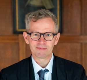 Portrait image of Tim Whitmarsh standing in front of a wood paneled wall wearing dark glasses and a dark blue suit and tie