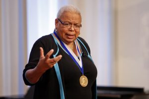 Image of Frances Smith Foster speaking to crowd and gesturing. She wears a black shirt and a medal around her neck.
