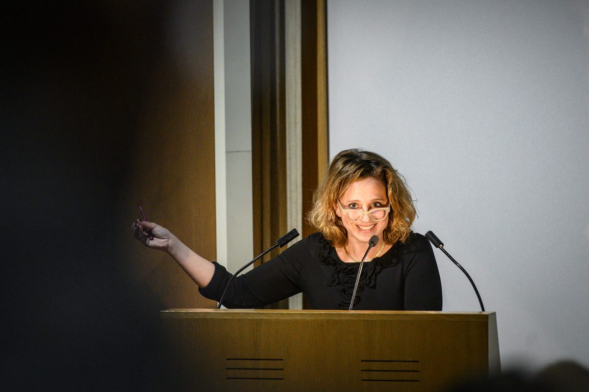 Photograph of a woman at a podium wearing a black shirt and glasses leaning over to speak into a microphone and pointing to the left (speaker's right)