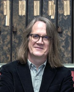 a photograph of Paul Taylor standing in front of shelves supporting filing boxes. The image is cut off at his chest. He is looking straight at the camera, and is smiling. He is clean-shaven and has long blond hair which rests on his shoulders. He is wearing glasses, a green and white striped shirt and a blue corduroy jacket. Behind him there are two rows of tall filing boxes, one red, the other mottled black and brown. Hand-written and typed labels on each box refer to their contents. The boxes contain photographs of works of art, and form part of the Photographic Collection of the Warburg Institute, where Paul works.