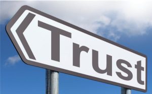 In this image there is a white road sign that says "Trust" on it and points toward the left top corner of the image. The background is a blue sky with clouds.