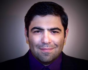 Color photograph portrait of a smiling man with dark hair in very dark contrast. Man wears a black suit with a purple shirt.