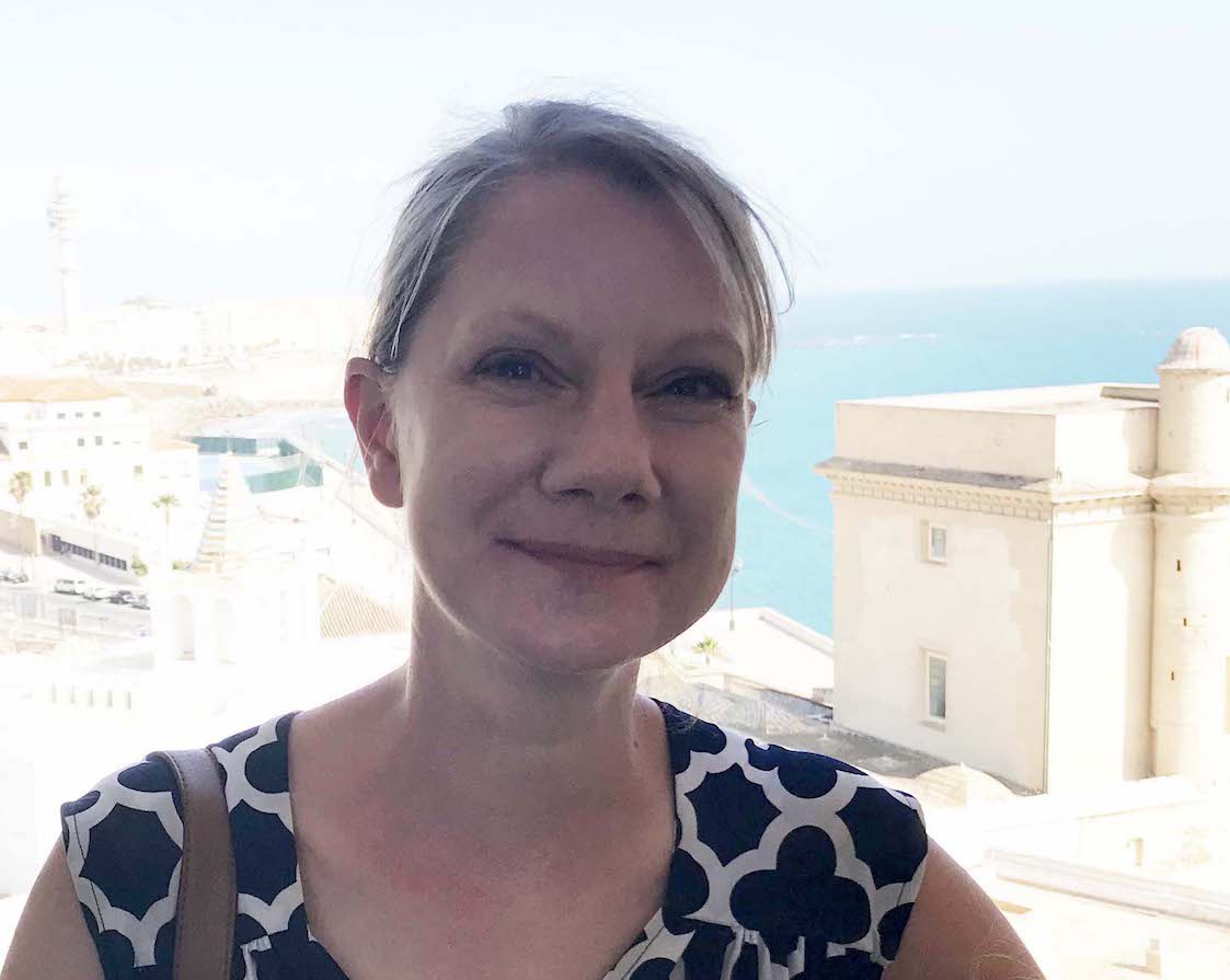 Photograph of Tanya Tiffany (white woman wearing a black and white dress with her hair pulled back) standing outdoors in Cadiz, Spain in front of a blue ocean.