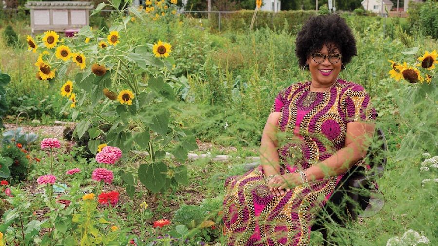 In this image Monica White sits smiling in a chair in a garden of plants and pink and yellow flowers.