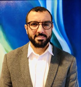 Image description: A bio photo for Baligh Ben Taleb. He is wearing glasses. He has on a tweed jacket with a white shirt underneath.