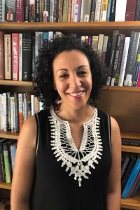 This is a picture of Laila Amine. She is looking directly at the camera and smiling. She is standing in front of a tall case of books.