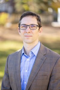 Image description: this is a headshot of Patrick Iber. He is sitting outside. He is looking directly at the camera and smiling.