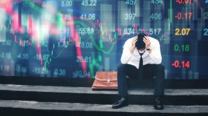A trader sits on the curb in front of a screen with images of the stock market crashing. His briefcase is next to him and his head is in his hands. 