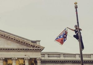 An African American woman, Bree Newsome, clings to the top of a flagpole. In her extended right hand is a Confederate flag she has just removed. In the background is the roof of the South Carolina statehouse.