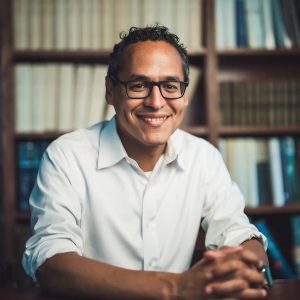 Image of a man in glasses and a white shirt seated at a desk in a library smiling.