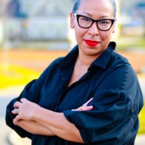Image description: This is an image of Shanna Greene Benjamin. She is standing up looking straight at the camera. She has on a black shirt and her arms are crossed across her chest. She is wearing black glasses.