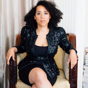This is an image of Caroline Randall Williams. She is sitting in an armchair with her legs crossed. She is looking directly at the camera. She is wearing a black dress with a black jacket over it.