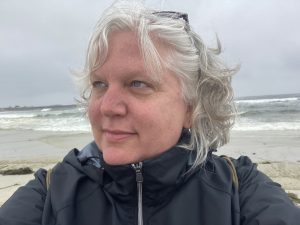 Headshot of a gray-haired white woman standing in front of the ocean in a black windbreaker, looking off camera