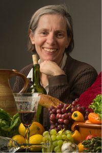 Headshot of Carolyn Nadeau, a smiling, white women with graying hair, her chin is leaning on her knuckles. In front of her are food images of a baguette loaf, grapes, peppers, lemons, garlic, asparagus and lettuce along with a large terracotta water pitcher, two glasses, one half-filled with red wine and another on its side, and a bottle of wine.