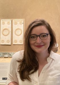 This is a headshot of a person with long brown hair standing in front of a museum display case. The display case contains two ivory panels of the consular diptych of Justinian (The Met, acc. nos. 17.190.52–.53).