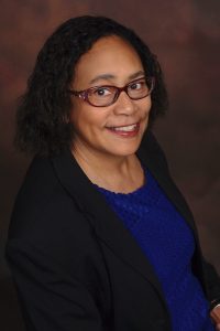 Womanwith glasses inblack jacket and dark blue dress.