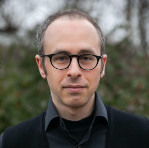 This headshot is of a man with short brown hair standing in front of a blurred background.