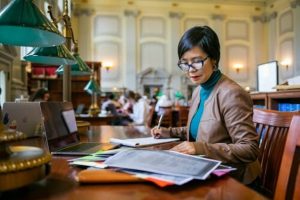 photo of woman wearing a brown jacket and glasses sitting in a library working.