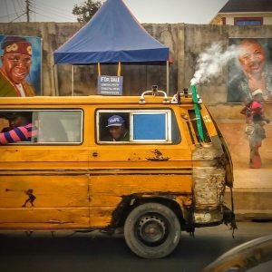 A rusty yellow bus, fitted with a PVC pipe for expelling exhaust, conveys passengers in Lagos, as a woman looks on; paintings of Nigeria's President Bola Tinubu and chair of the Nigerian Union of Road Transport Workers overlook the road.