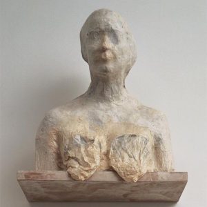A faceless person’s torso with deflated breasts rests on top of a shelf over a white background. It’s made of white paper and wood. 