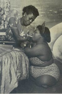 In the image above from a 1951 issue of Ebony magazine, a Black woman with short hair, sideshow performer Pearl Jeffries is perched atop a bed and leans in closely to light the cigarette of fellow performer Alzora Green, pictured here as a Black woman wearing a crown.