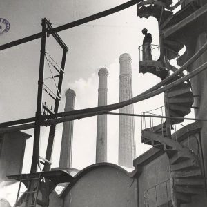A figure stands atop a spiral staircase overlooking the Lafarge factory in Casablanca, the plant's three smokestacks in the background.