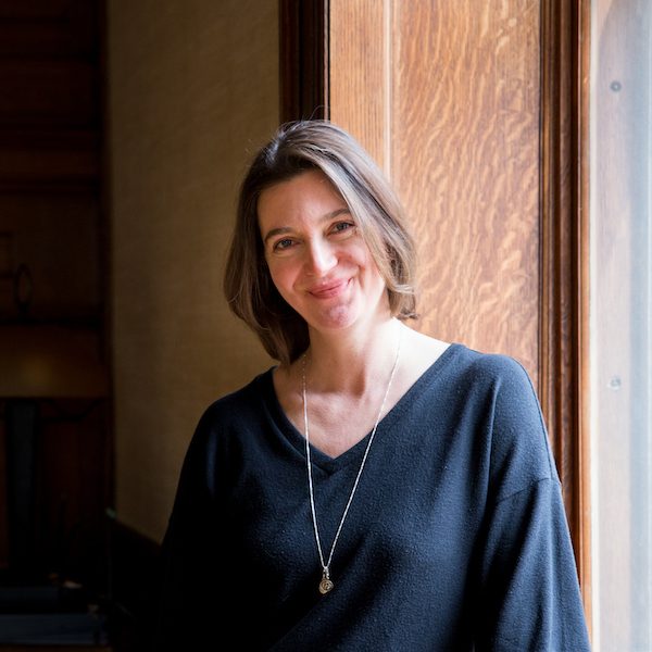 This is a photo from the waist up of a smiling woman with shoulder-length brown hair, wearing a black sweater, and leaning against a wide, dark wood windowsill with sunlight coming in.
