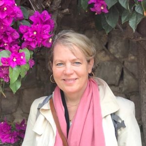 This is a headshot of a woman with blonde hair standing outside next to flowers.