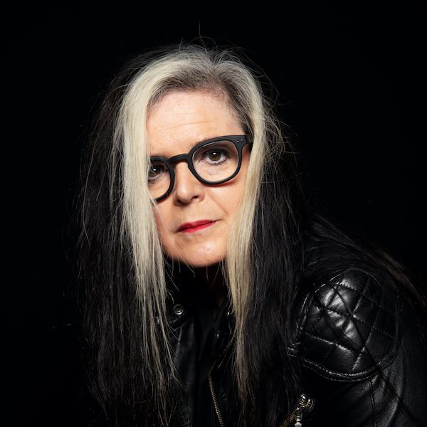 A color headshot composed as a study in noir, the photograph illuminates the quilting on the shoulder of Casid’s well-worn black leather jacket, the contrast of their hair, jet except for its two face-framing flashes of white, and their dark-eyed, pale face in a field of black with raven spectacles accentuating the intensity of their gaze.