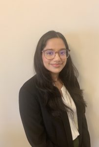 This is a headshot of a woman with long black hair and brown glasses standing in front of a white wall.