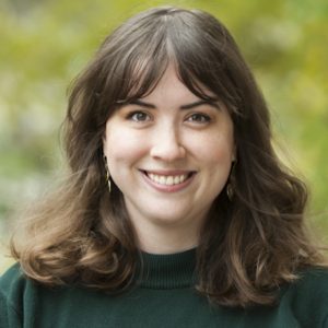 This is a headshot of a woman with shoulder-length brown hair standing outside with greenery behind her.