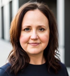 This is a headshot of a woman with shoulder-length brown hair standing outside with greenery behind her.