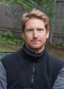 man with blonde hair wearing a black vest stands in front of greenery