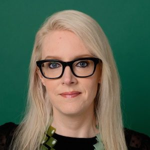 This is a headshot of a woman with long blond hair and black-framed glasses in front of green background.