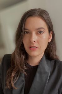 woman wearing grey blazer and black shirt looks directly at the camera. She has brown hair and brown eyes.