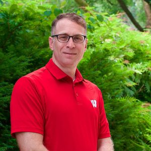 Dan Kapust stands in front of greenery. He is wearing black glasses and a red UW-branded polo shirt.