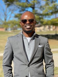 Tolu stands in front of a tree. He wears a grey suit and sunglasses.