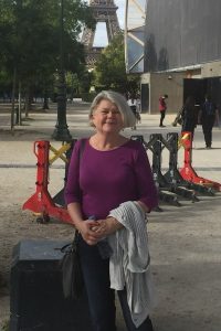 Anne Vila stands in front of the Eiffel Tower. She is wearing a purple shirt and is holding a jacket.