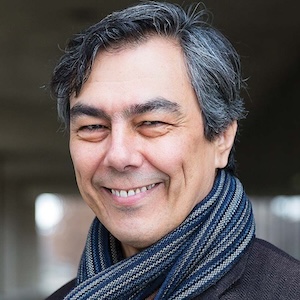 Mario Ortiz-Robles smiles at the camera. He is wearing a scarf and has black-grey hair. 