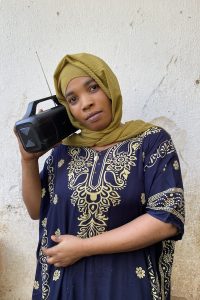 A woman wearing an olive-green hijab and a navy-blue dress with gold floral embroidery holds a portable radio to her ear, gazing directly at the camera.