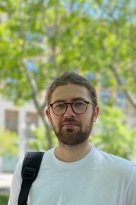 A man stands in front of a tree. He has brown hair and facial hair and is wearing black glasses and a white t-shirt. He has a backpack on.