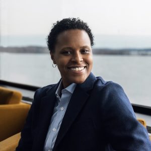 Image of a woman in front of water. She has short curly dark hair and is wearing a dark blue blazer and light blue button up shirt and hoop earrings. She is smiling.
