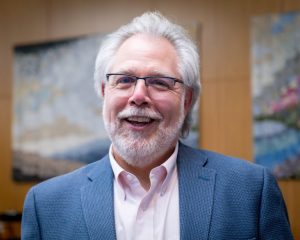 Photograph of a man indoors with white-grey hair and glasses wearing a blue blazer and pink shirt, and smiling. 