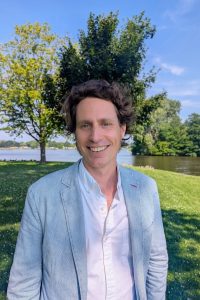 This is a head-and-torso shot of a man with brown hair standing outside with trees behind him.