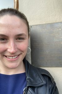 Image of a woman wearing a leather jacket and blue shirt standing in front of a plaque about Rene Descartes