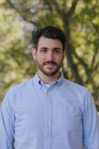 This is a headshot of a man with short brown hair standing outside with greenery behind him.